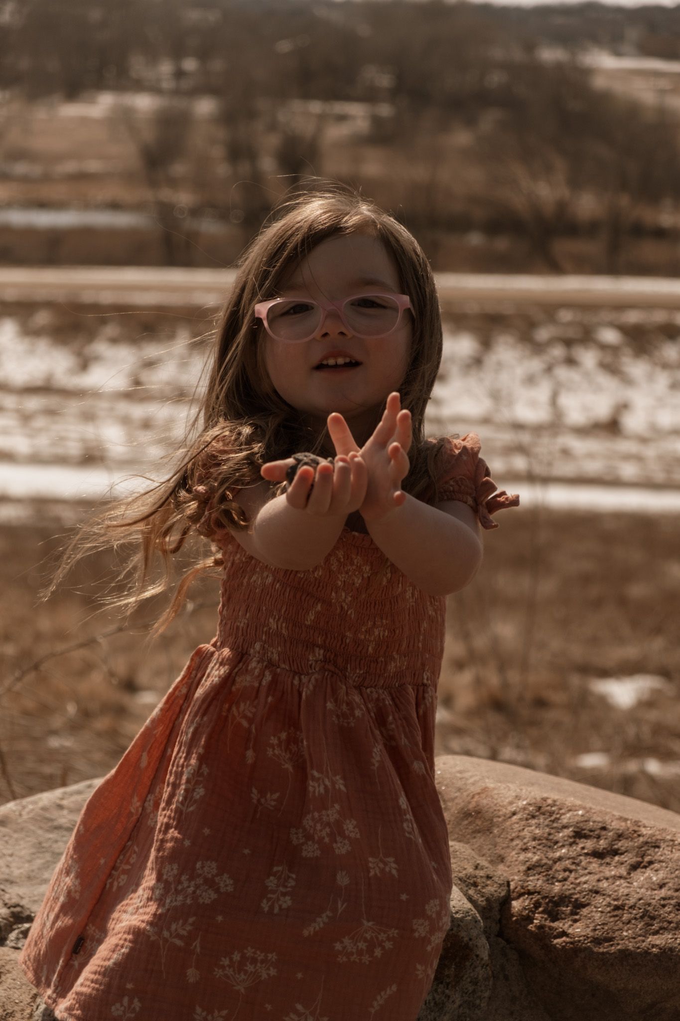 A small child in a floral dress plays outdoors in warm sepia lighting on a rocky landscape.