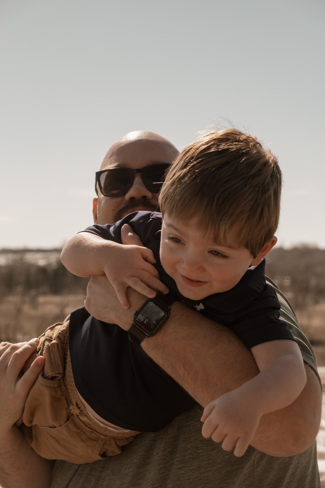 A child in brown overalls smiles while being playfully held by someone wearing sunglasses during a sunset.
