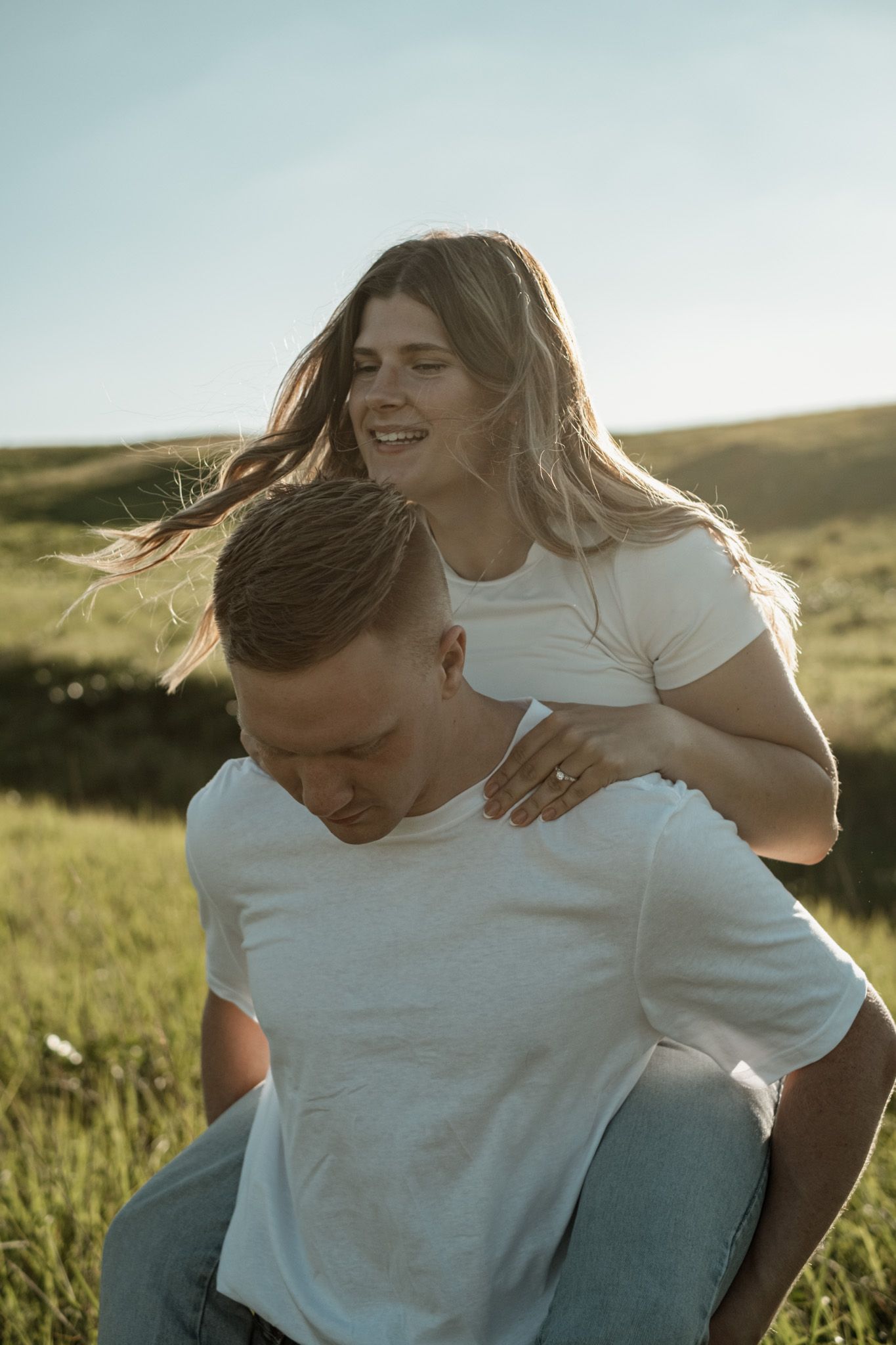A couple wearing white t-shirts embraces and laughs together in a sun-drenched meadow.