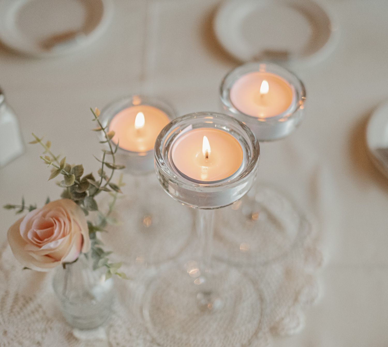 Lit white tealight candles in glass holders with delicate pink roses and eucalyptus on a lace tablecloth.