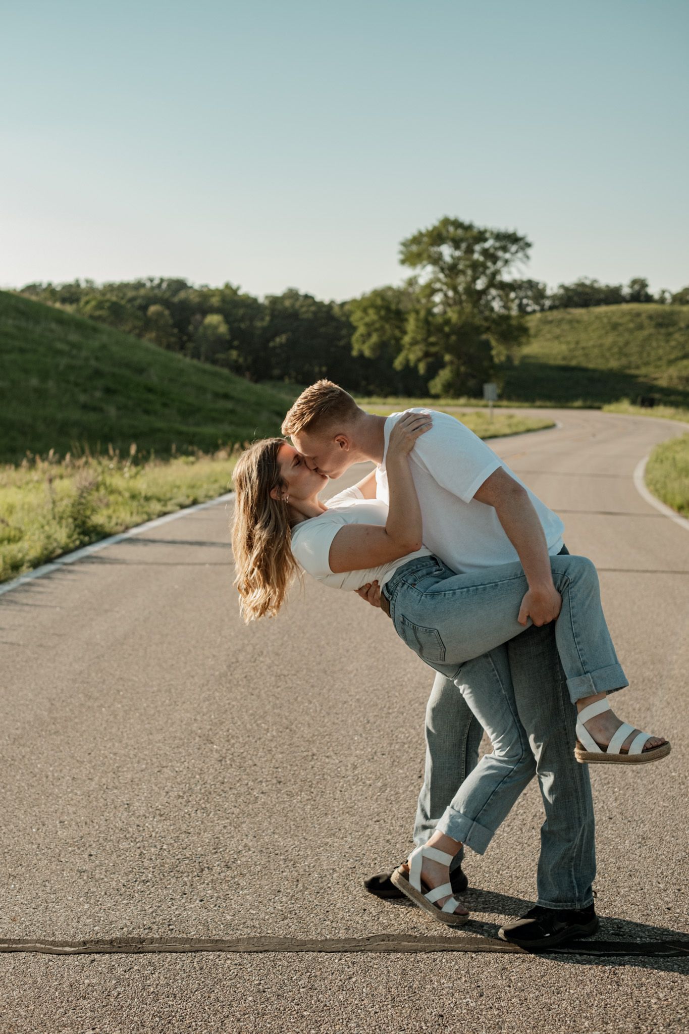 Playful couple dances and dips on an empty country road surrounded by green rolling hills.