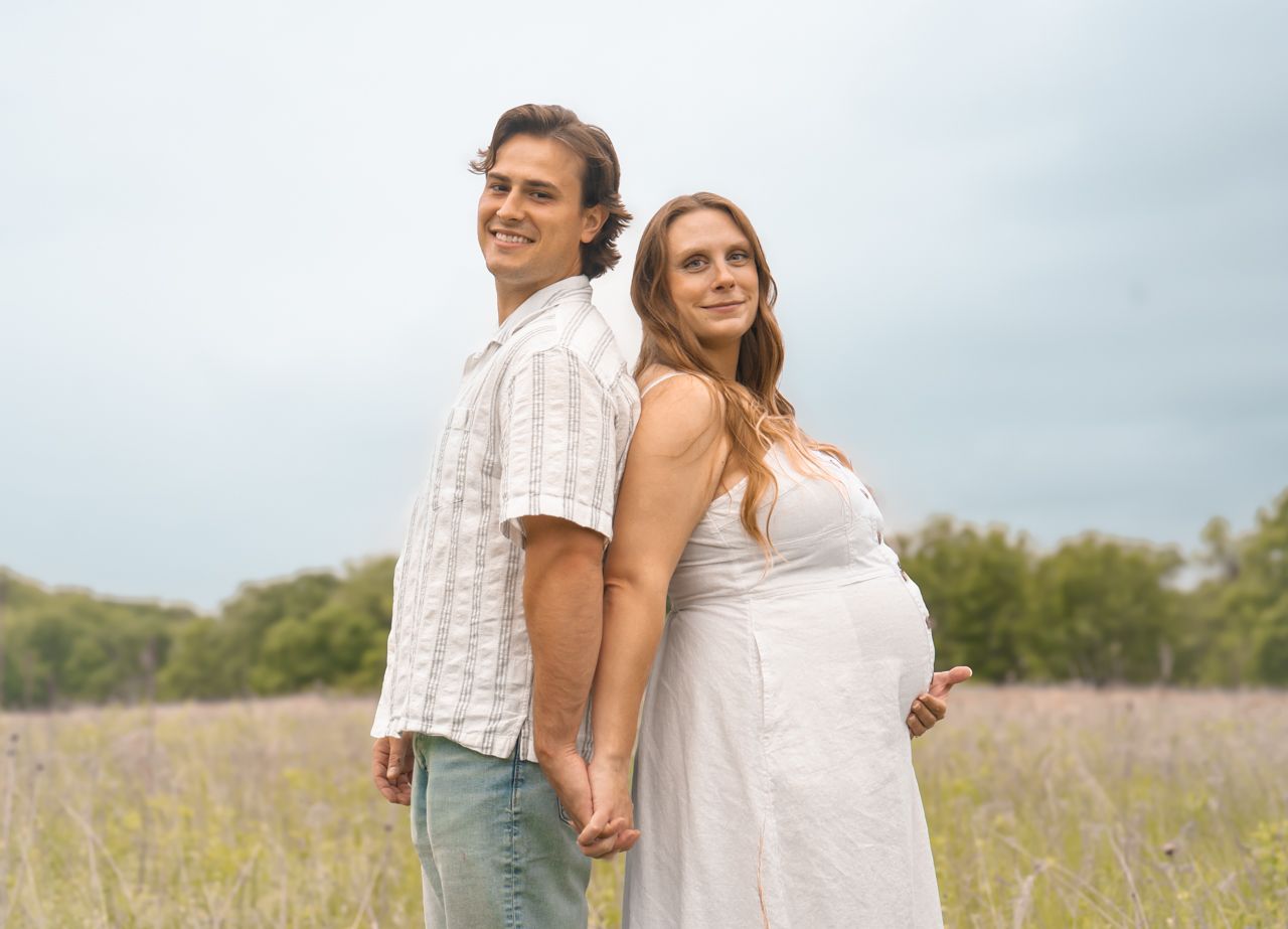 Expecting couple poses back-to-back in a sunlit field, with her wearing a white dress and him in casual attire.