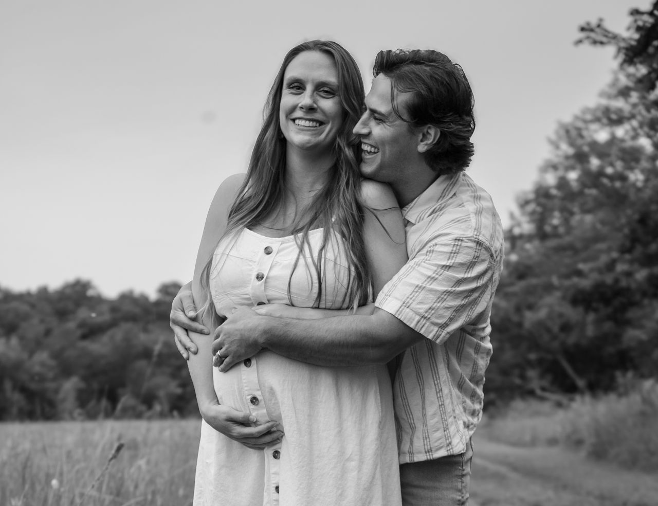 A romantic black and white portrait of an expectant couple embracing in a sunny field.