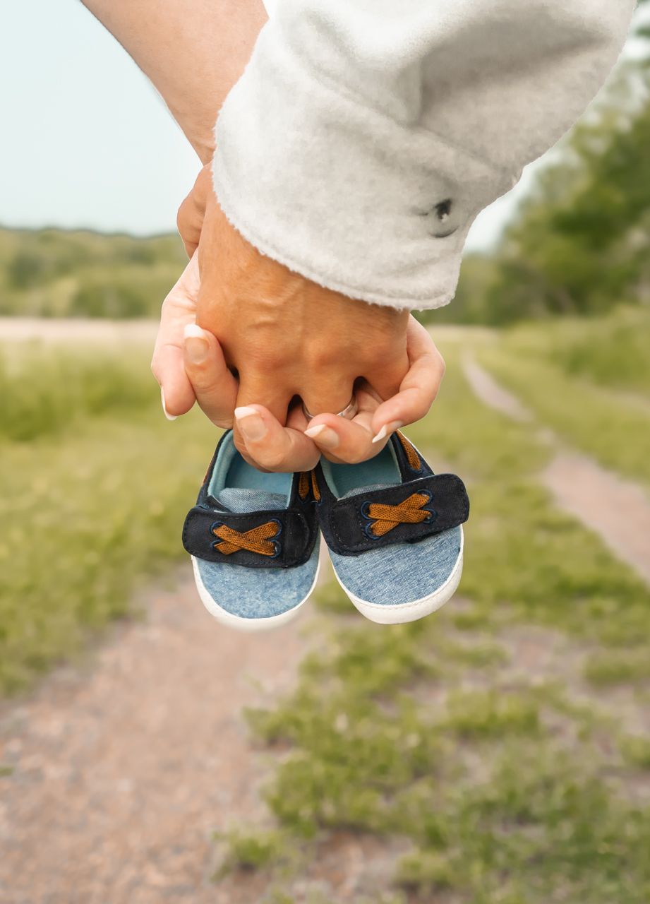 Hand holding tiny blue baby shoes on a dirt path with green grass in the background.