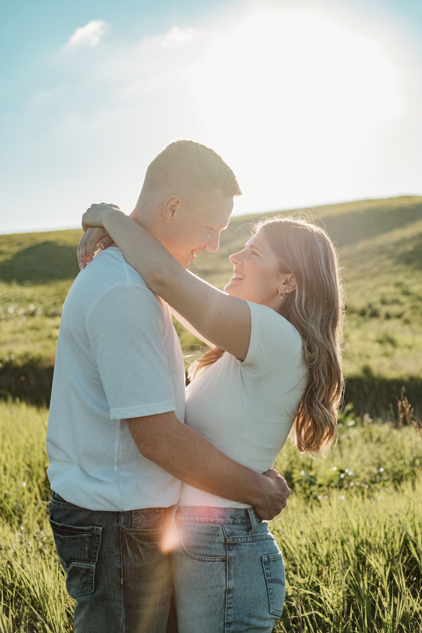 Romantic couple embraces in a sunlit meadow during golden hour, dressed in white and denim.