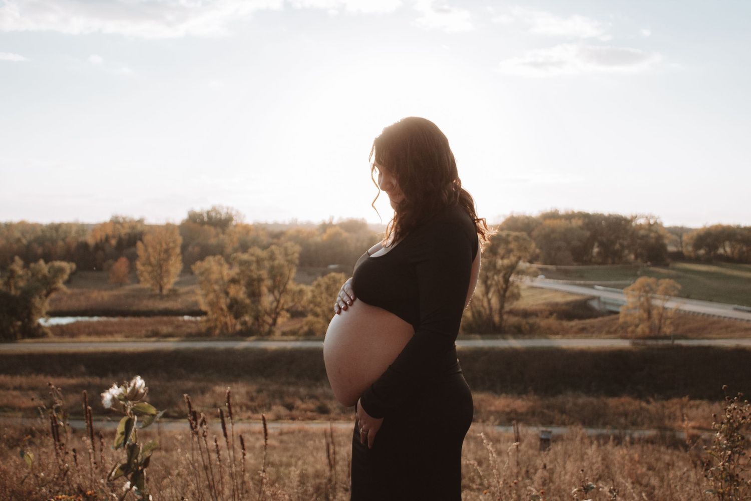 A silhouetted pregnant woman stands in front of golden autumn landscape during sunset while holding her belly.