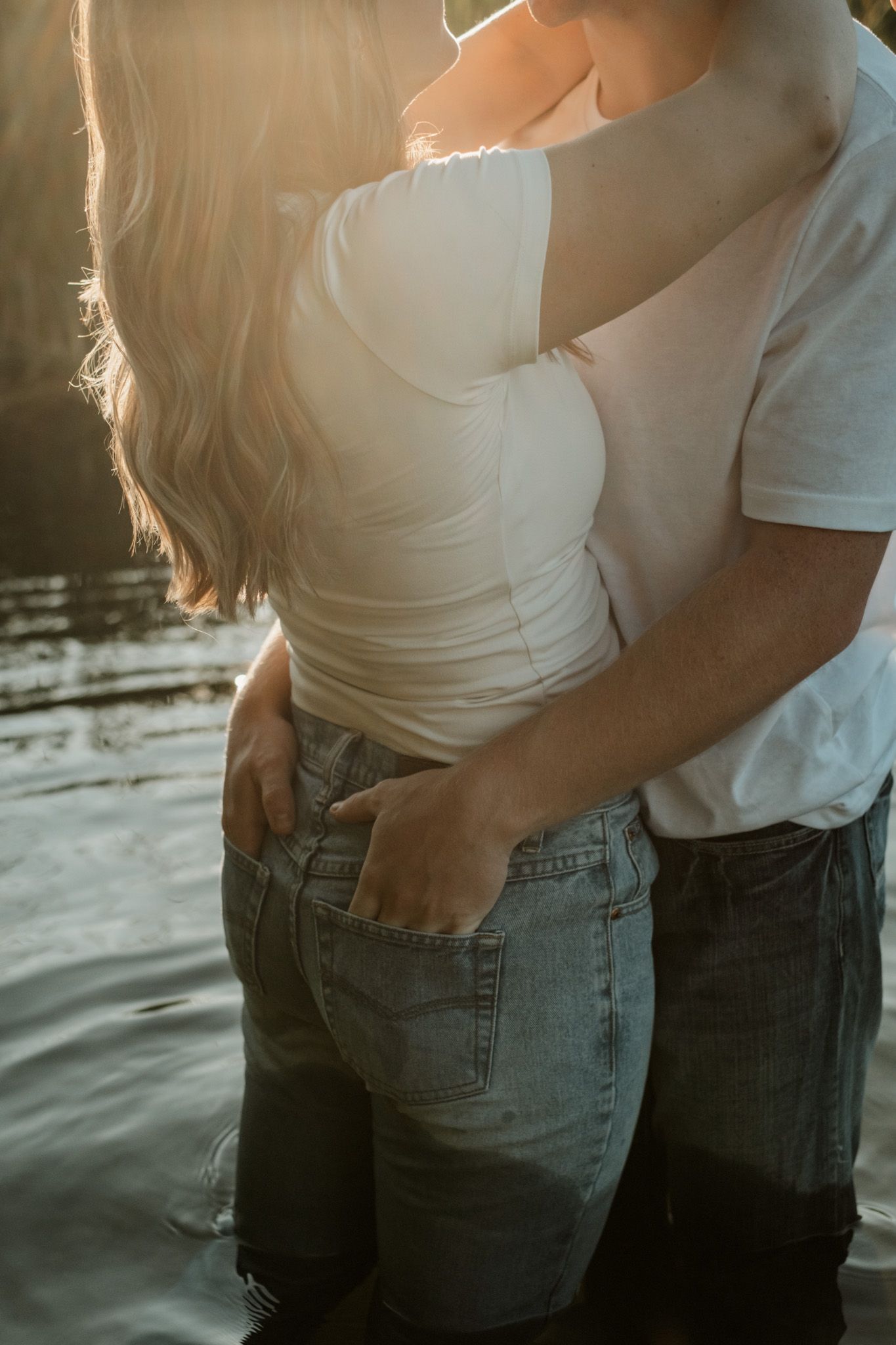Romantic couple in casual white tops and jeans embracing by the water at sunset with golden sunlight.