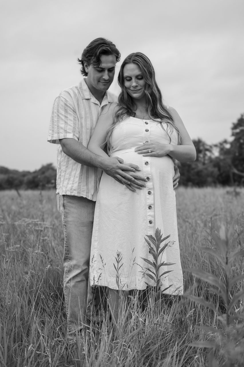 Black and white portrait of an intimate maternity moment in a field with a couple in casual summer attire.
