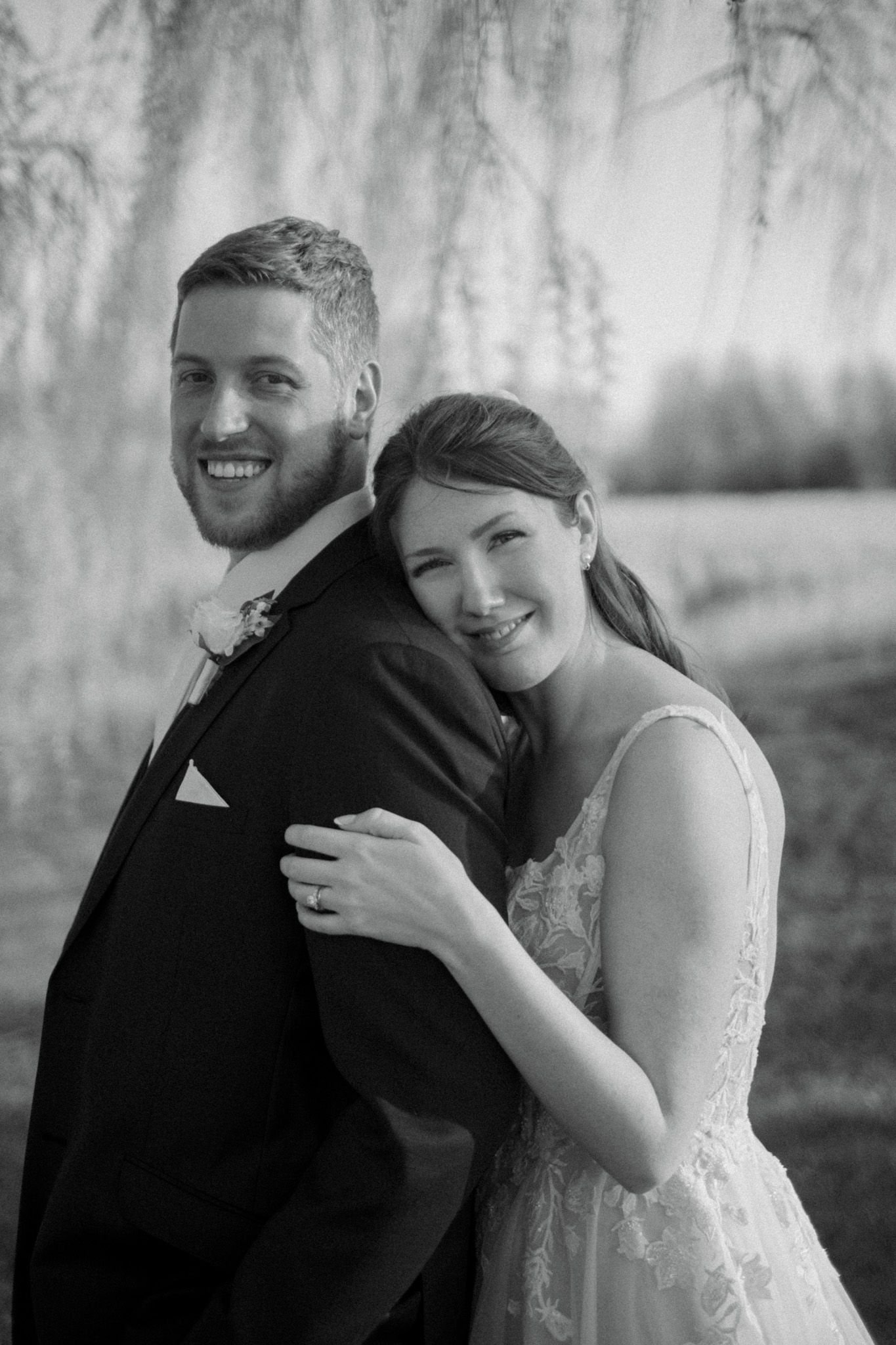 A black and white wedding portrait shows a couple embracing under willow trees in a romantic outdoor setting.