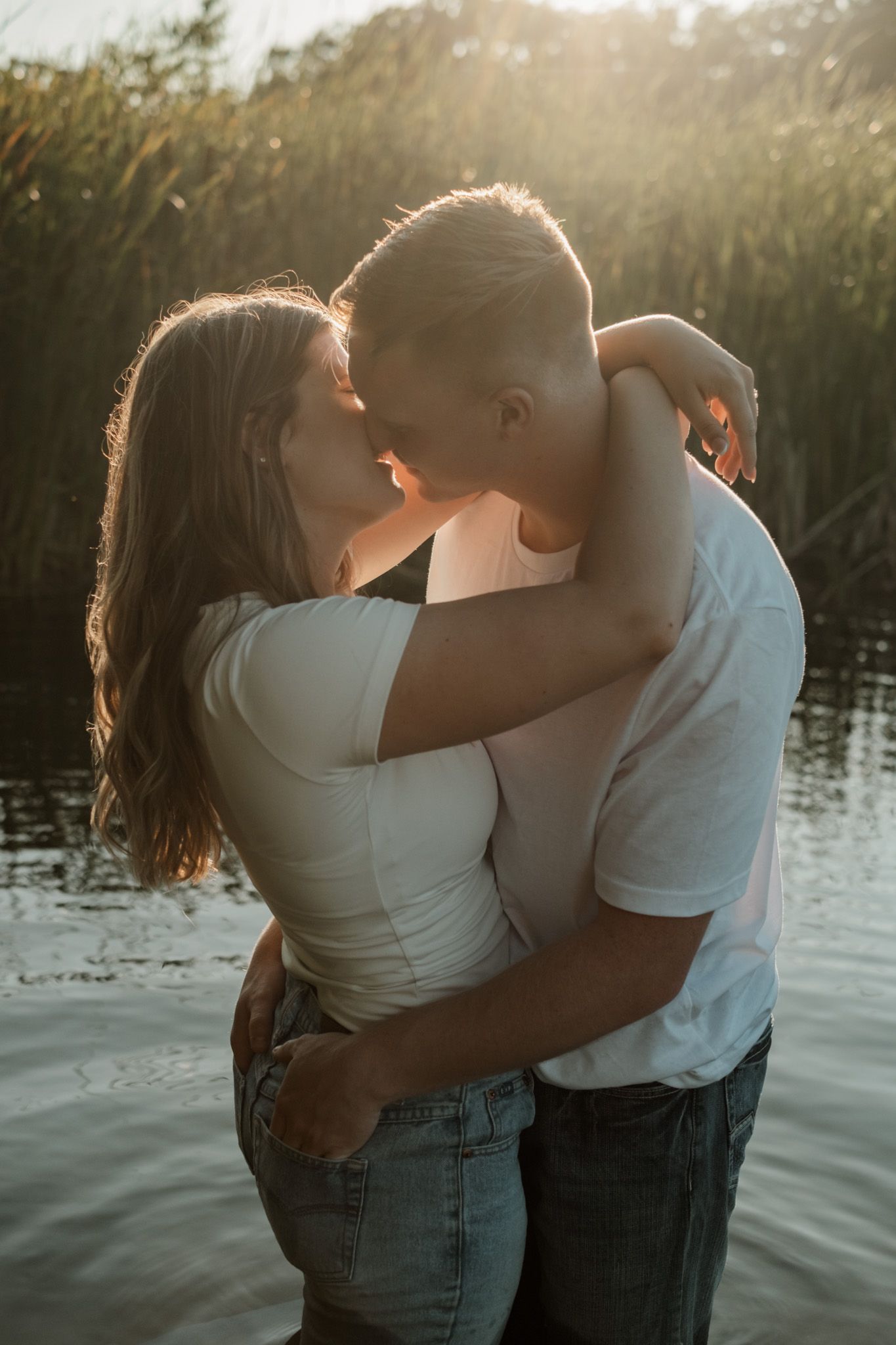 A romantic couple embraces and kisses by a lake at sunset with tall grass and golden light surrounding them.