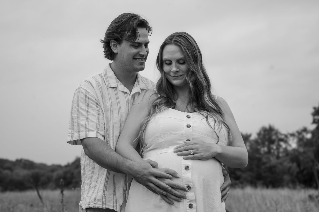 Black and white photo of an expectant couple embracing in a field while standing close together.
