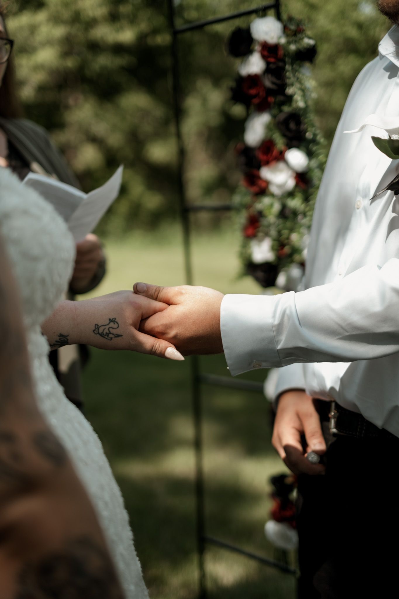 Hands in white sleeves hold each other during an intimate wedding ceremony moment under floral decorations.