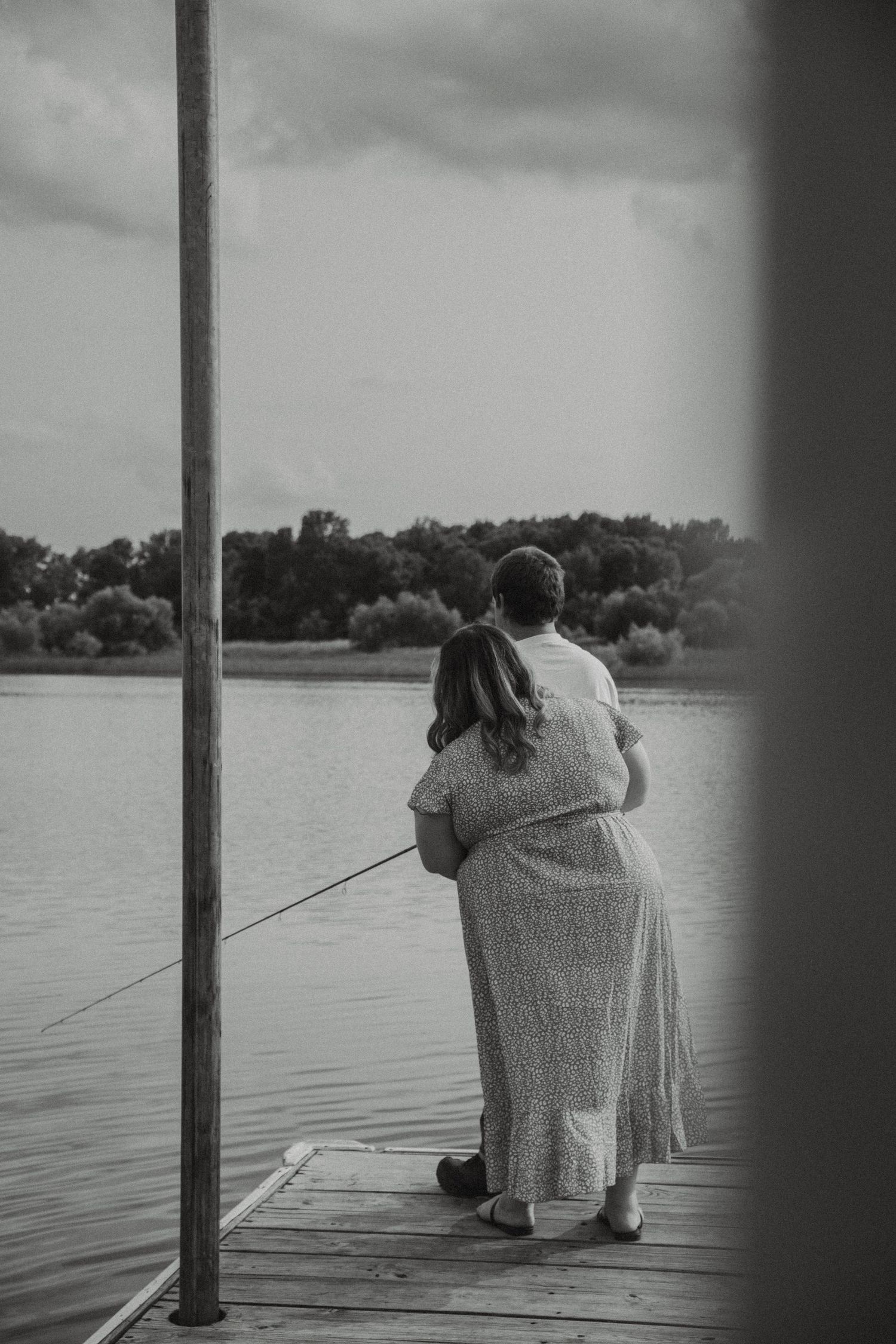 Black and white photo showing two people embracing while fishing from a wooden dock on a serene lake at dusk.