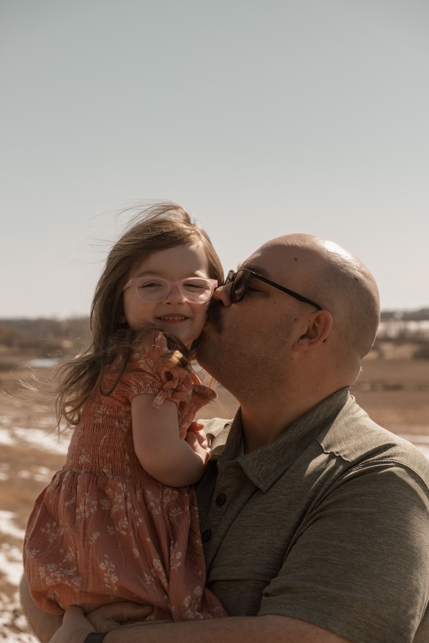 A tender moment captured outdoors with a close-up of an adult sharing affection with a young child in a coral dress.
