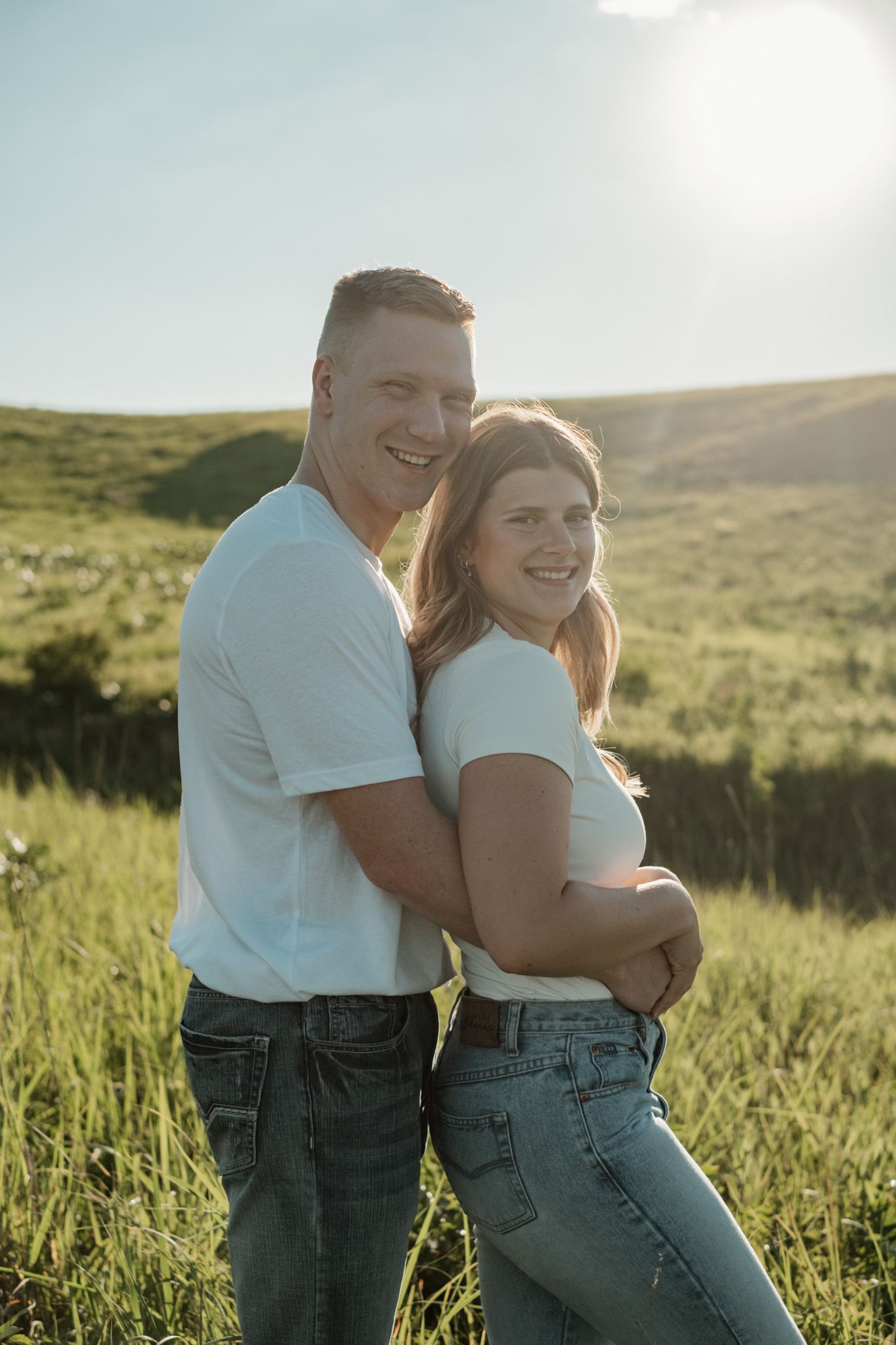 A couple in matching white shirts and jeans shares playful moments in a sunlit grassy field.