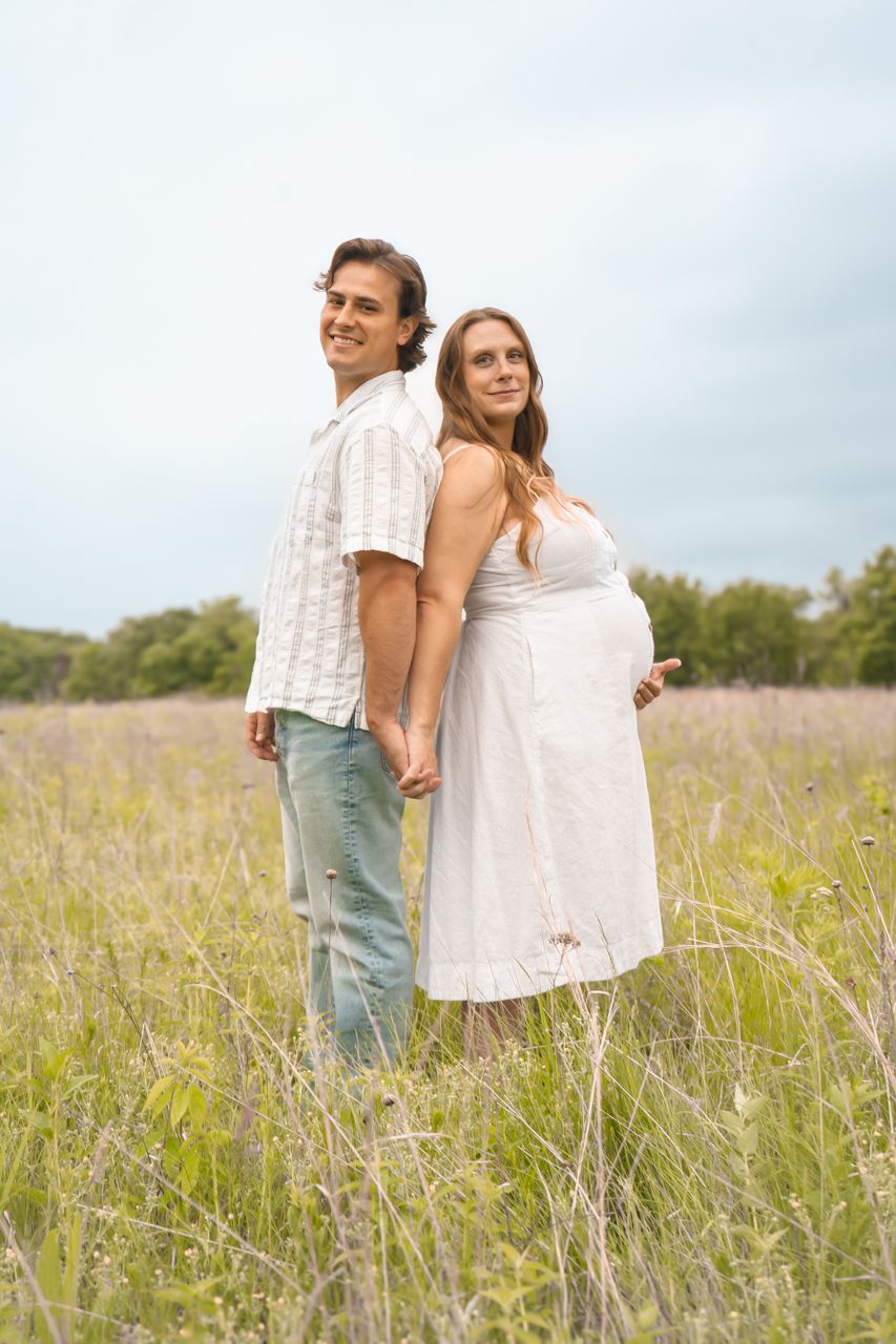 A couple stands together in a sunny meadow of tall grass, with the woman wearing a white dress.