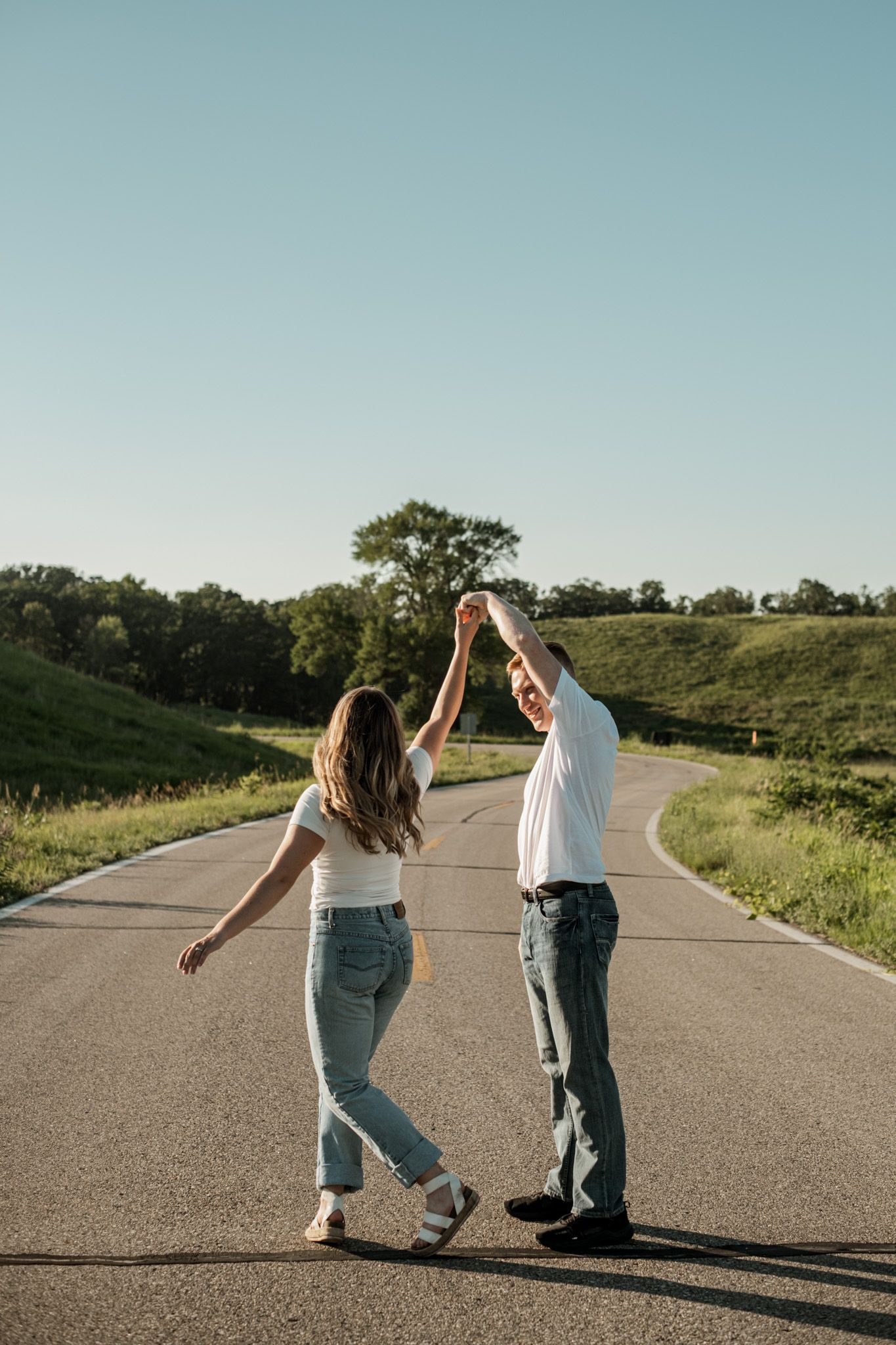 A couple dances and spins together on an empty rural road surrounded by green hills.