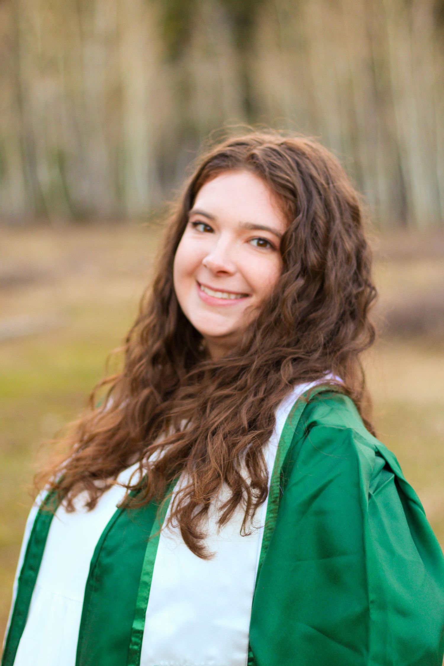 Smiling young woman with long curly brown hair wearing a green graduation gown outdoors among trees.