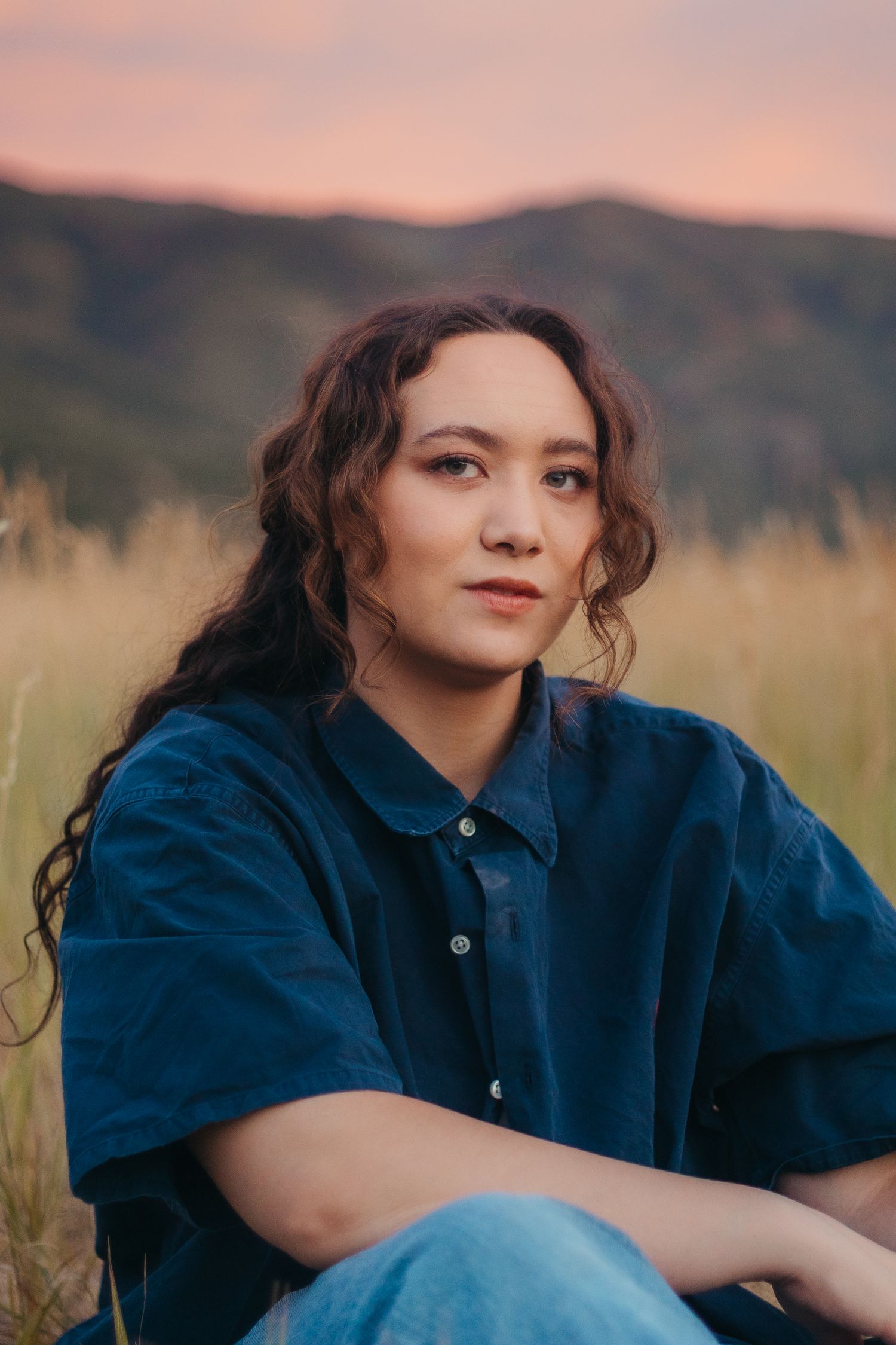 Young woman in oversized navy shirt sitting in golden grass field at sunset with mountains behind.