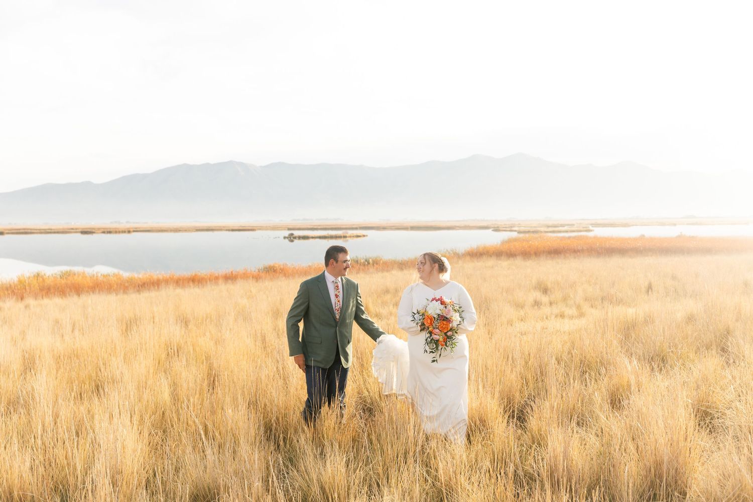 Bride and groom walk hand in hand through golden marsh grass with mountain lake backdrop in soft sunlight.