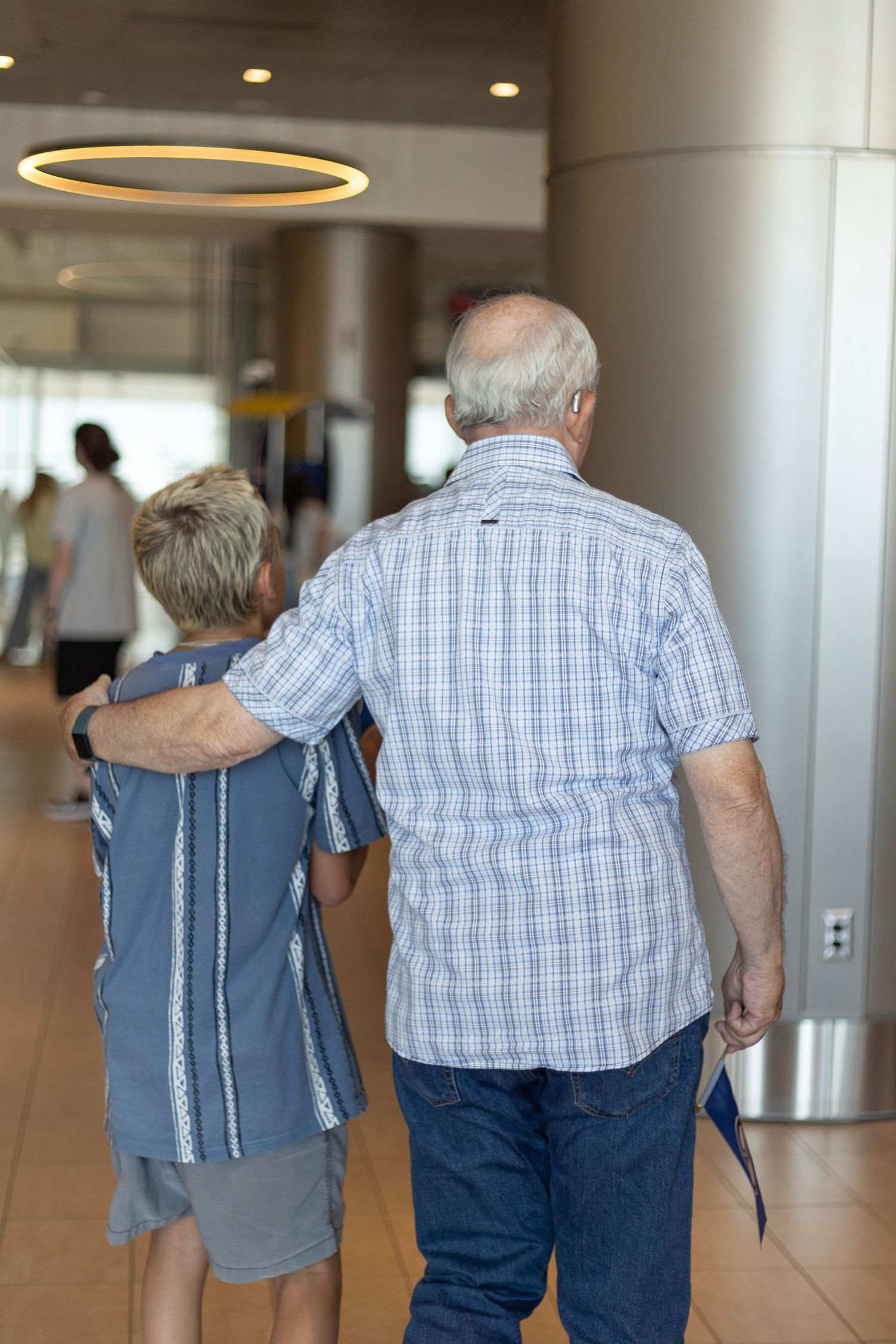 Elderly couple walking arm in arm down a modern building hallway, viewed from behind.