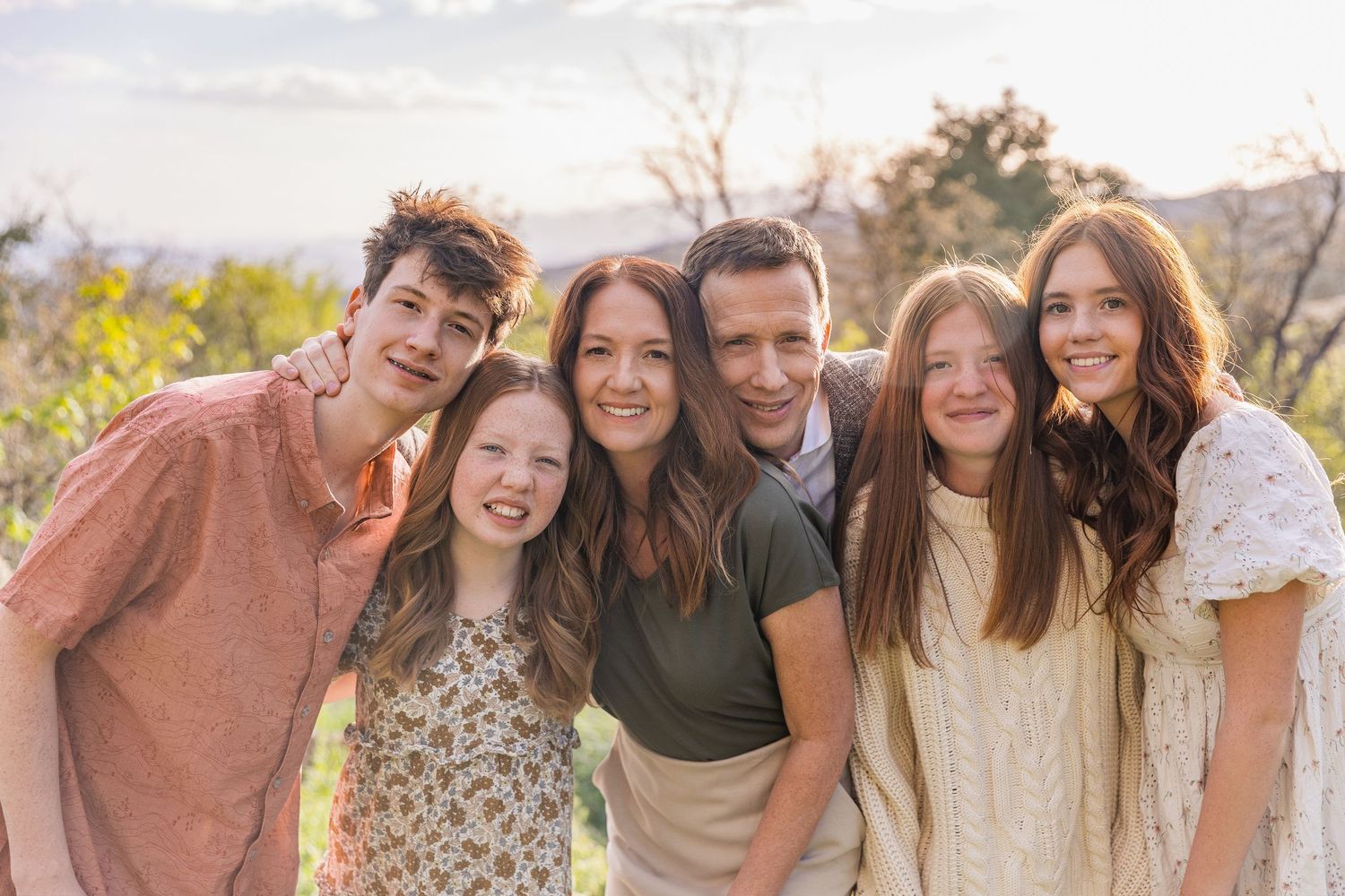 Happy family of six posing outdoors in warm sunlight with scenic mountain views in the background.