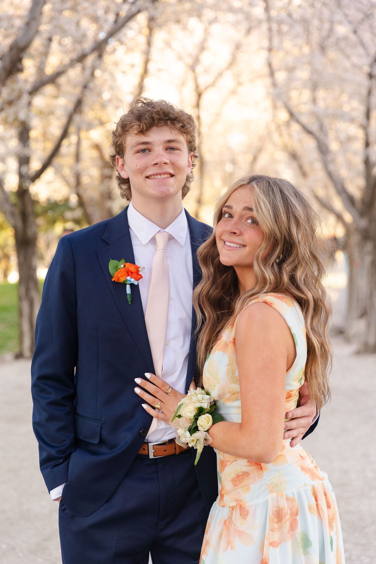 Smiling prom couple outdoors among cherry trees, boy in navy suit and girl in floral dress.