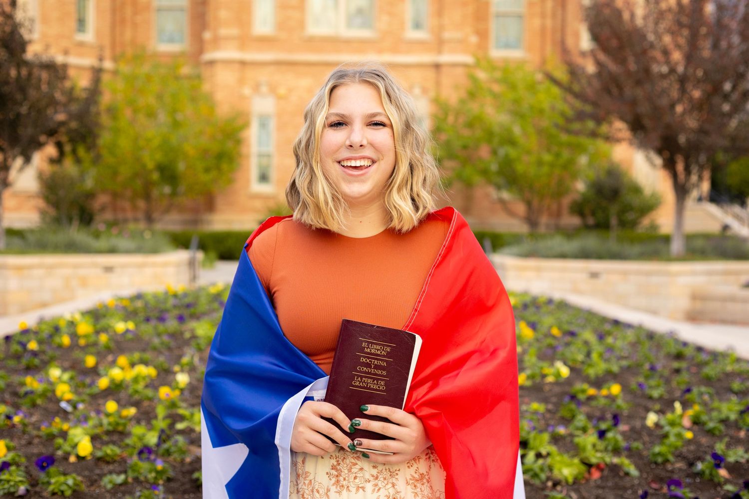 Smiling young woman wrapped in Chilean flag holding a book outside a brick building.