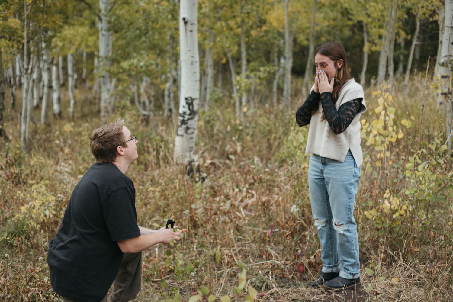 Man kneels to propose to emotional woman in an autumn aspen forest setting.