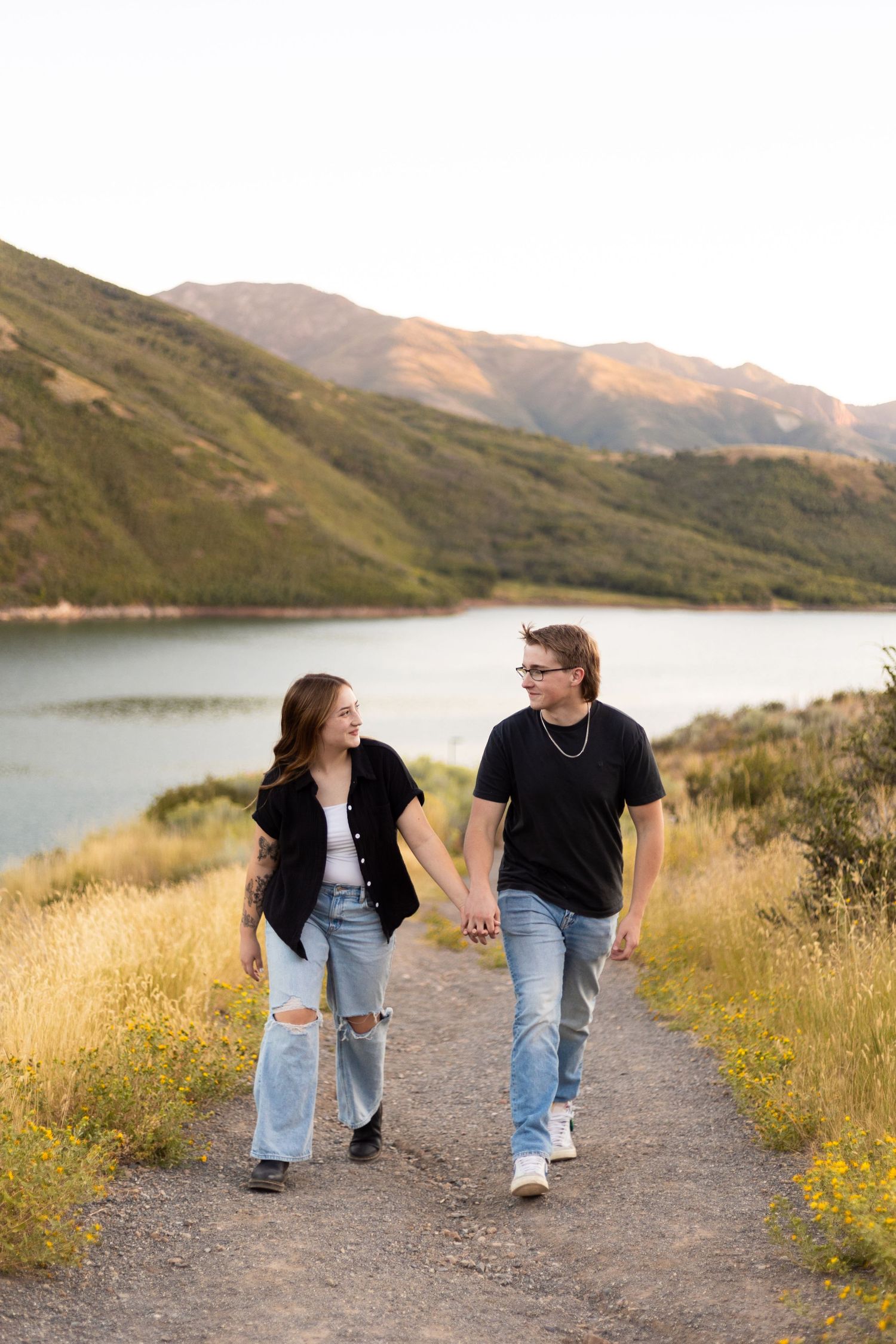 Young couple holding hands strolling along a scenic path with a mountain lake behind them.