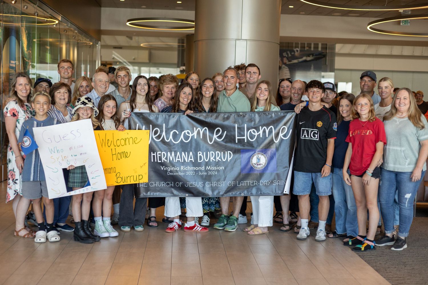 Large group of friends and family gather with Welcome Home Hermana Burrup banner at airport.