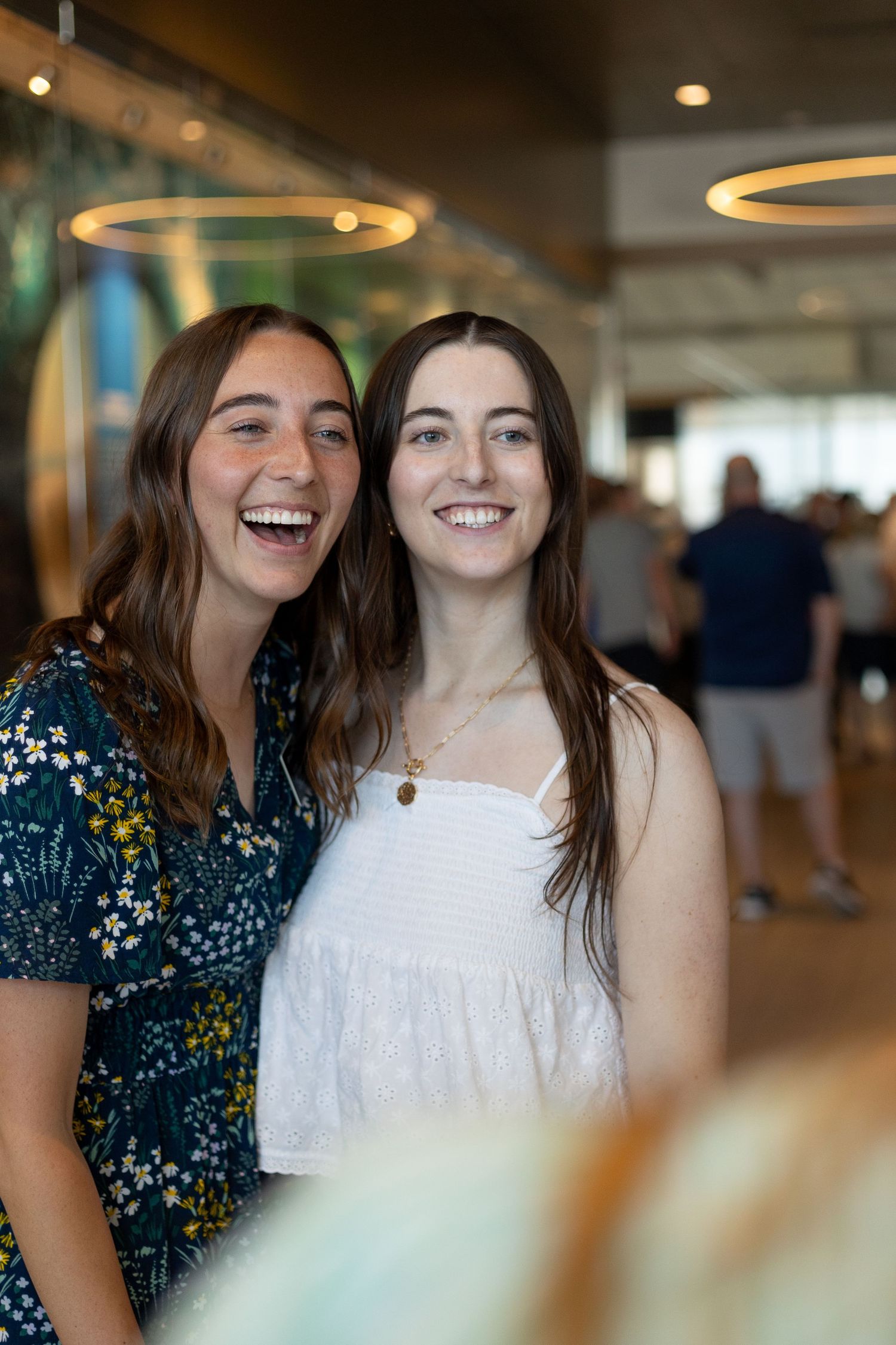 Two young brunette women laugh and smile together at a joyful homecoming celebration at the airport