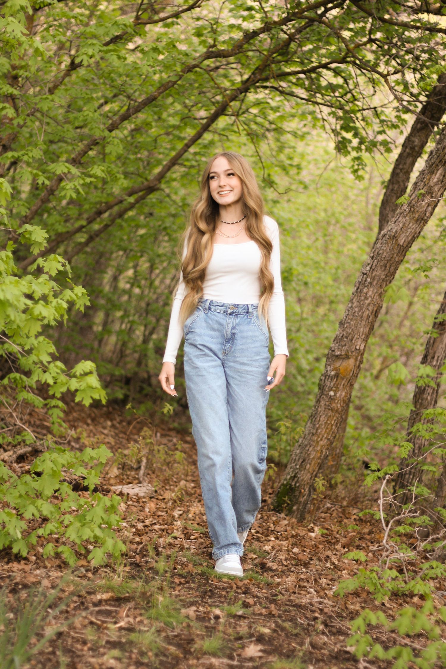 Young woman with long blonde hair in jeans and white top walking along a wooded forest trail in spring.