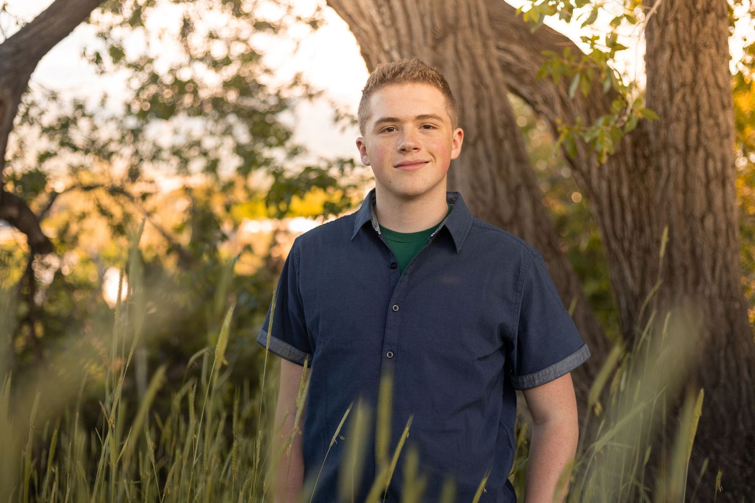 Smiling young man in navy shirt standing among tall grass near trees at golden hour.