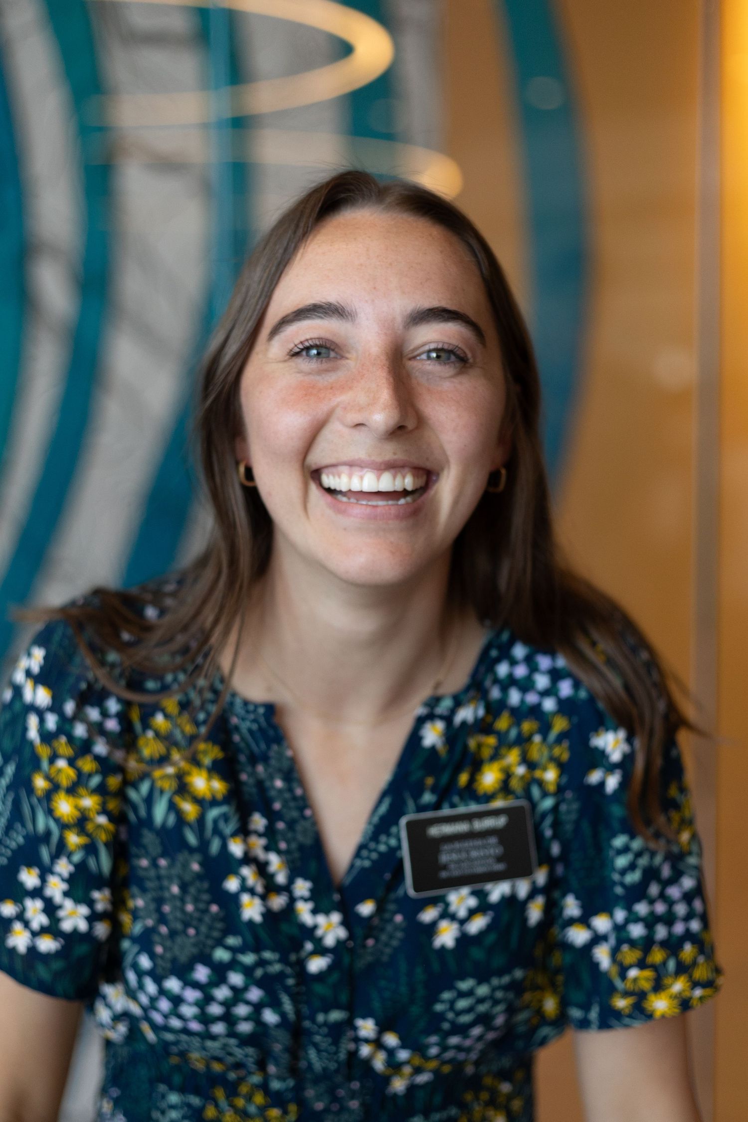 Joyful young woman in floral dress wearing missionary name tag smiles brightly at camera.