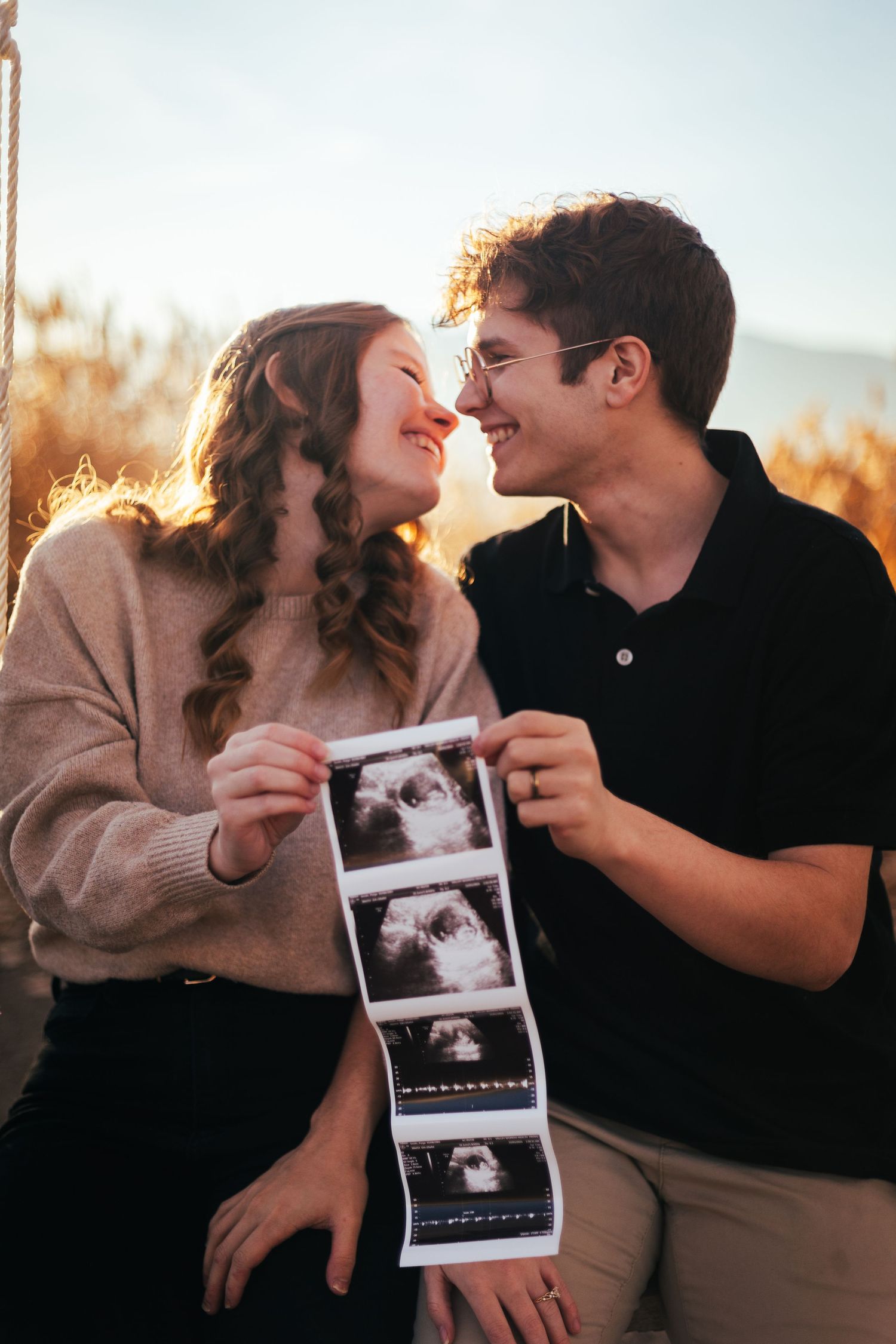 Smiling couple holds ultrasound photos close together in a warm sunlit outdoor autumn setting.