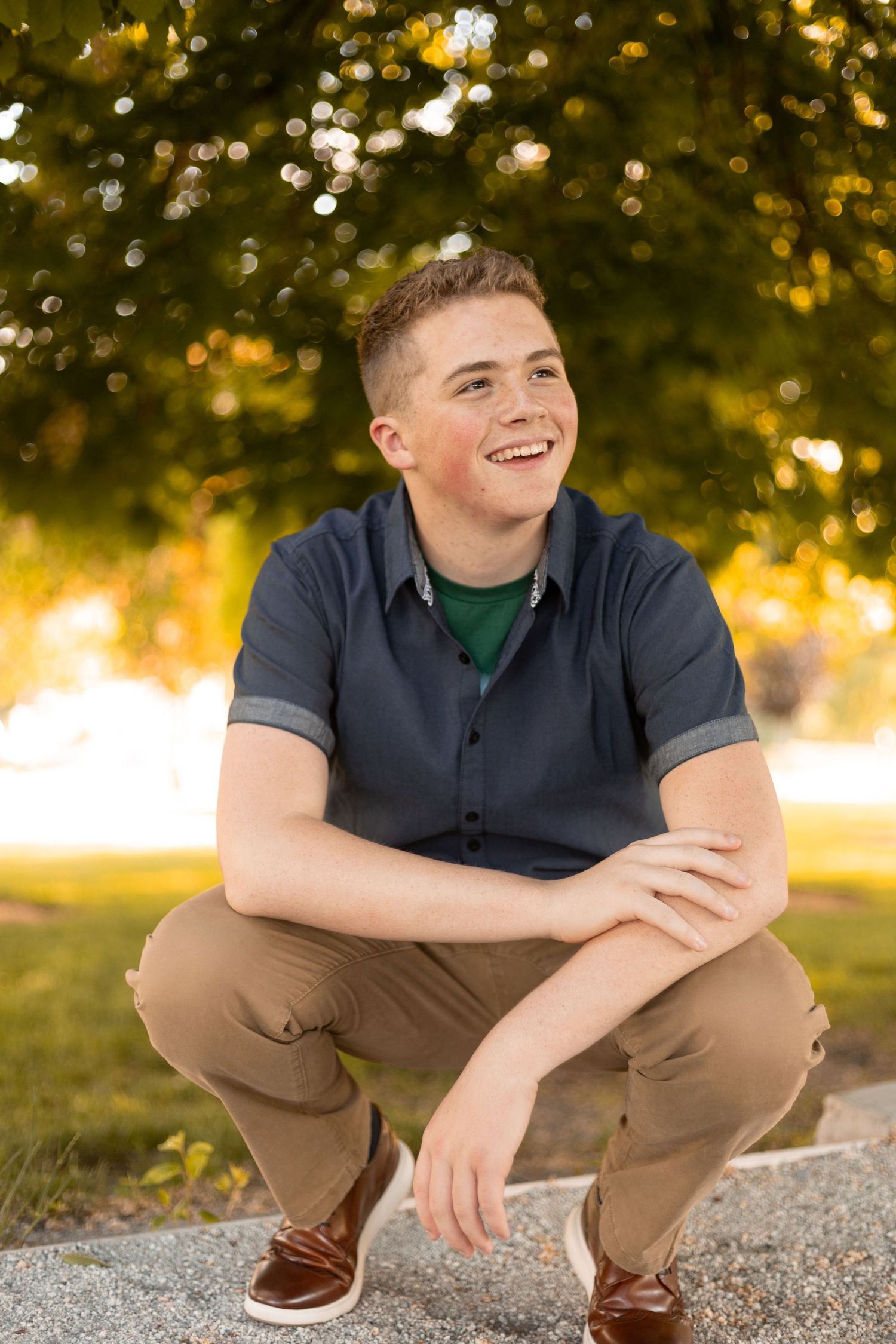 Happy teenage boy in a navy polo shirt and khaki pants crouching outdoors with green trees in the background.