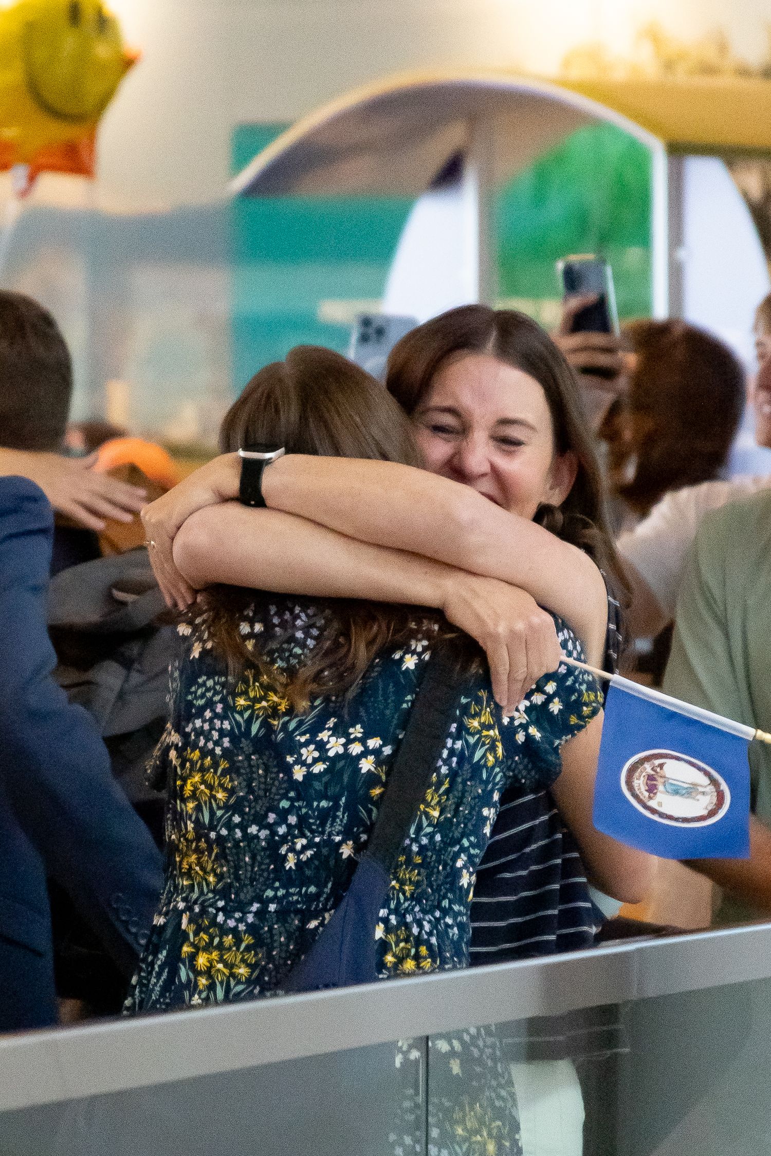Mom and return missionary daughter share a joyful embrace at an airport reunion while holding a small flag.