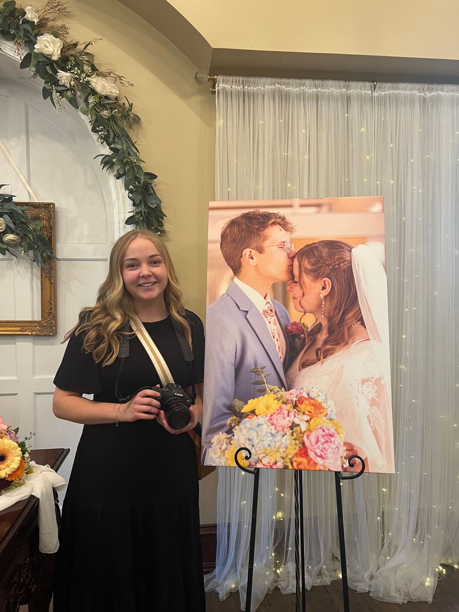 Kaylee Anne in black dress stands beside a large wedding photo display holding a camera.