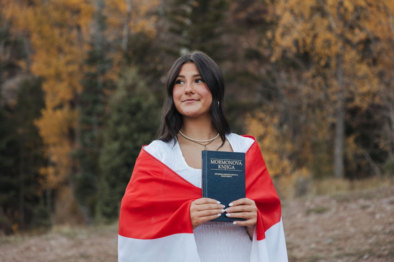 Future missionary draped in red and white flag holds a book outdoors among autumn yellow trees.