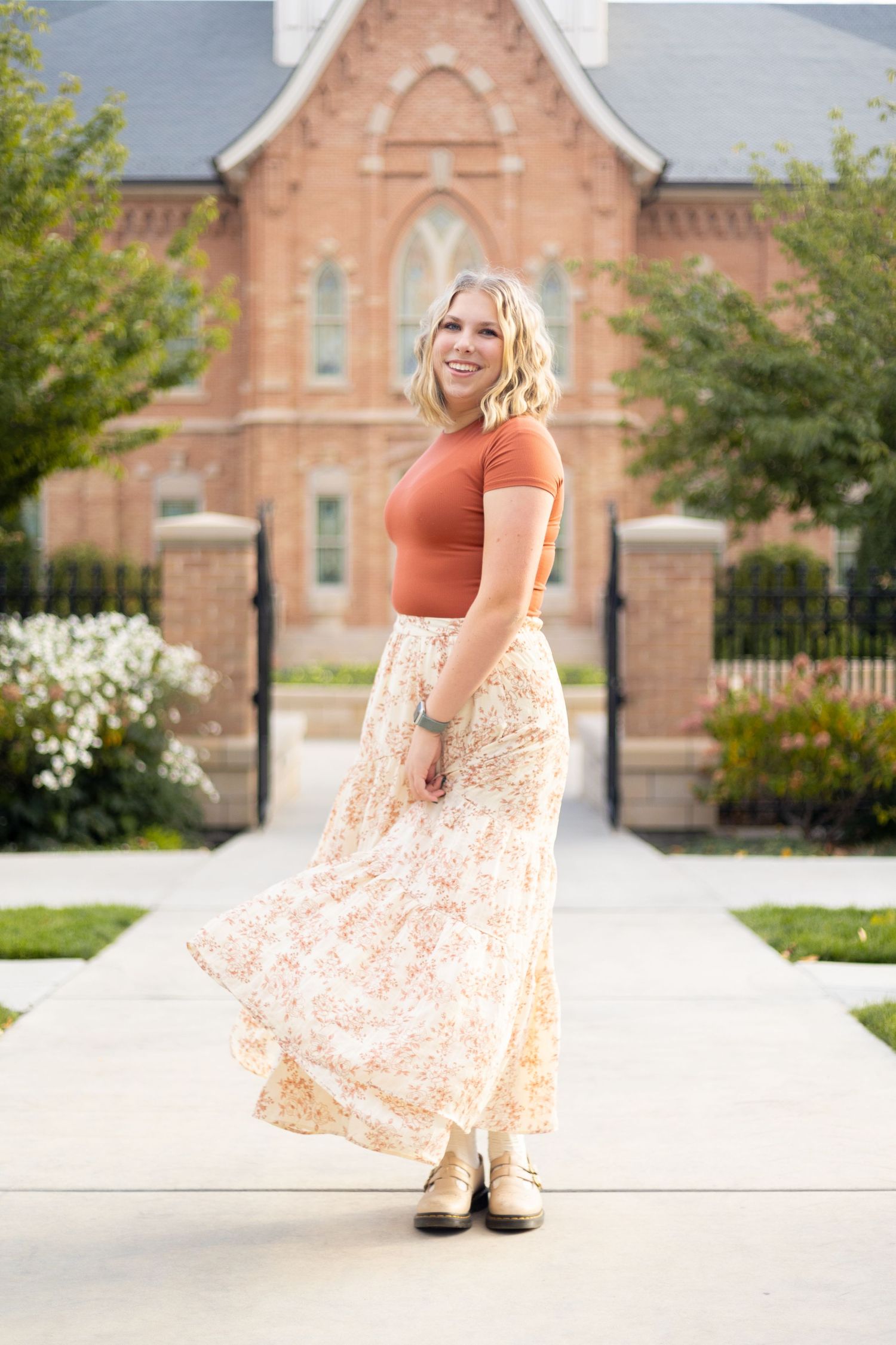 Blonde woman in rust top and floral maxi skirt twirling outside a brick church building.
