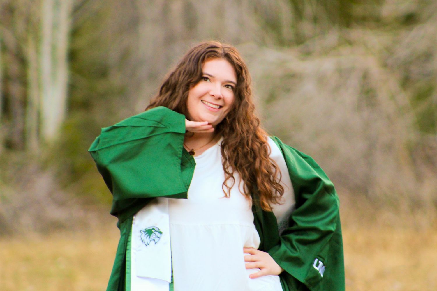 Smiling young woman in green graduation gown posing outdoors in natural wooded setting.
