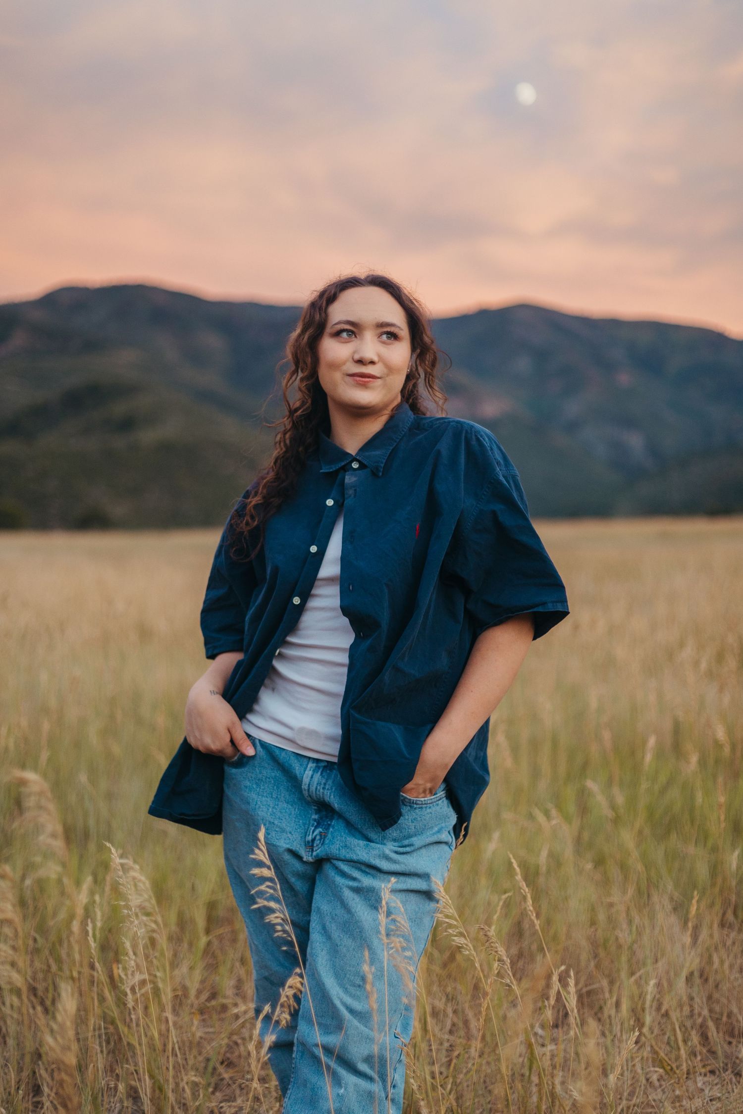 Young woman in navy shirt and jeans standing in golden field at dusk with mountain backdrop.