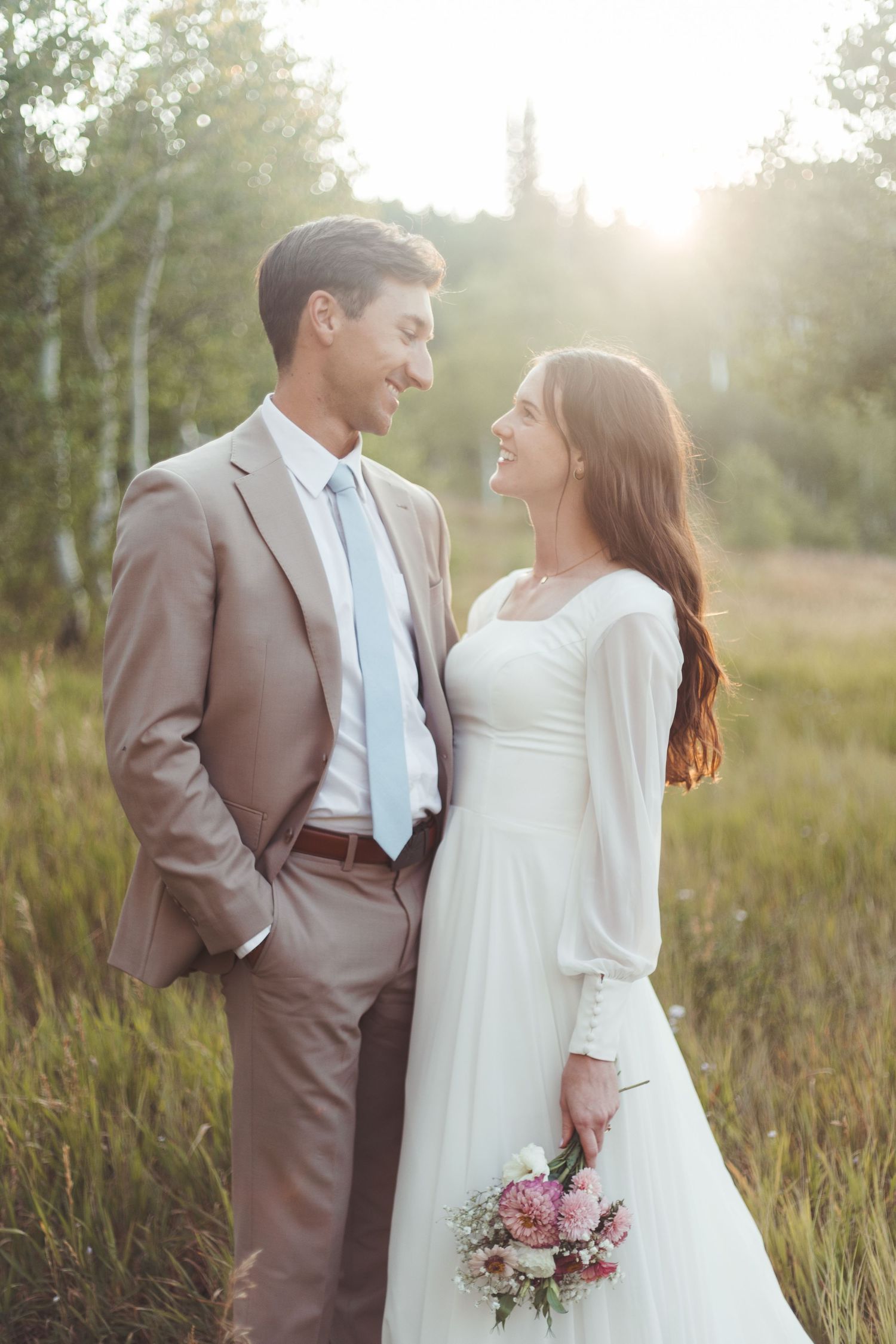 Happy bride and groom smile at each other in sun-drenched outdoor field, bride holding pink floral bouquet.