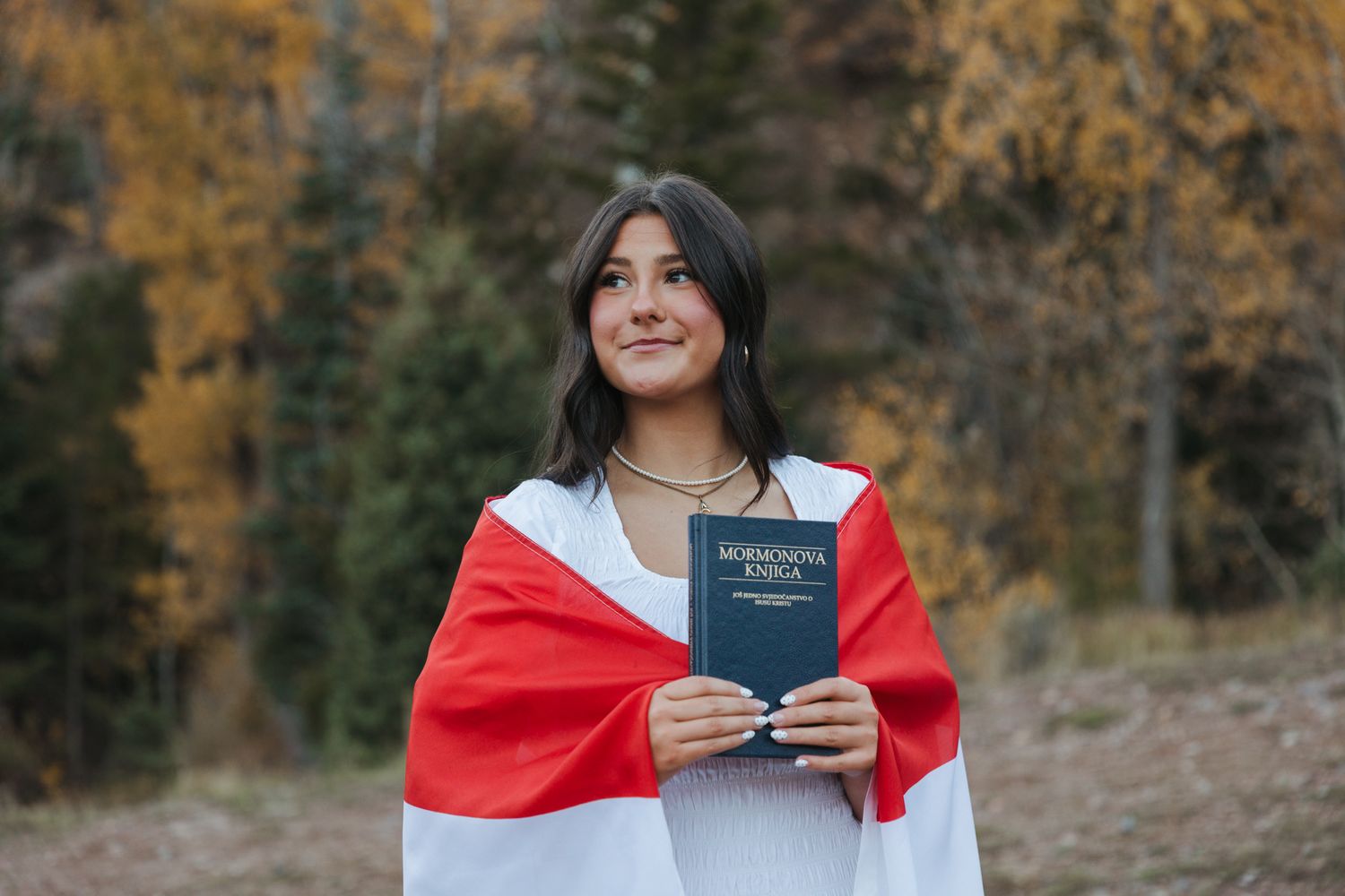Young woman wrapped in red and white flag holding a book in autumn forest, future missionary with a Book of Mormon.