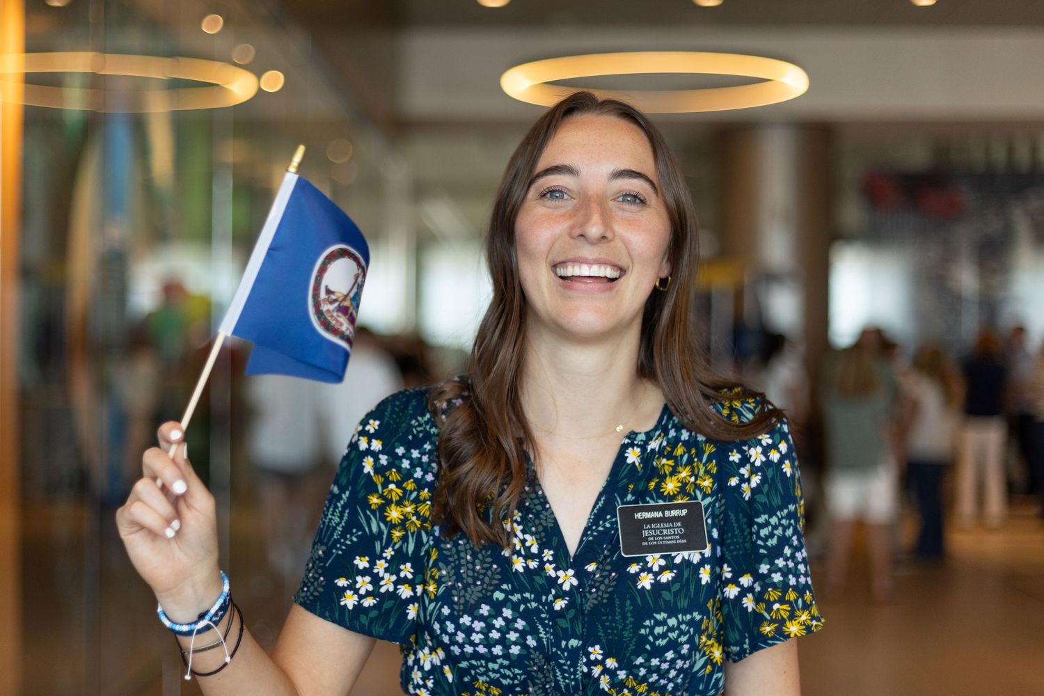 Smiling young woman in floral scrubs holding a Virginia state flag inside a bright building.