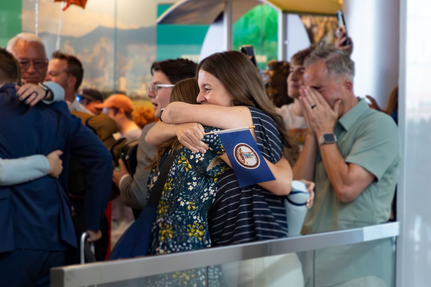 Mom & returned missionary daughter sharing an emotional hug at an airport arrival gate surrounded by other reuniting people.