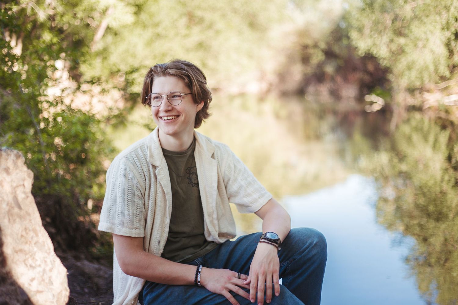 Teenage boy in glasses sitting by a reflective river, smiling in natural outdoor setting.