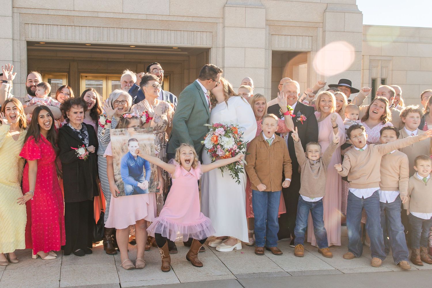 Large joyful wedding party group photo outside a temple with bride and groom kissing in the center.