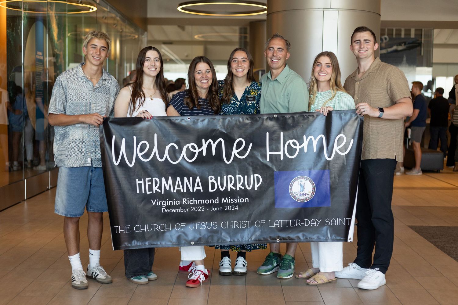 Family group holds Welcome Home Hermana Burrup Virginia Richmond Mission banner at airport arrival.